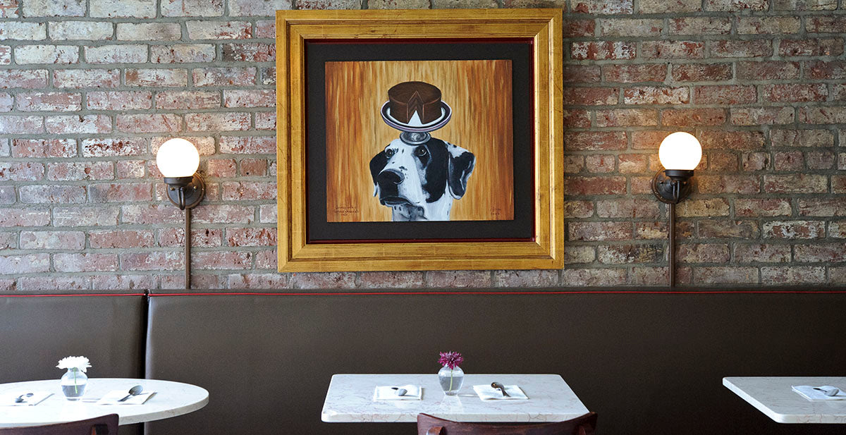3 marble tabletops service ready along a brown banquet,  brick wall with 2 sconces around a painting of a cake on a dogs head