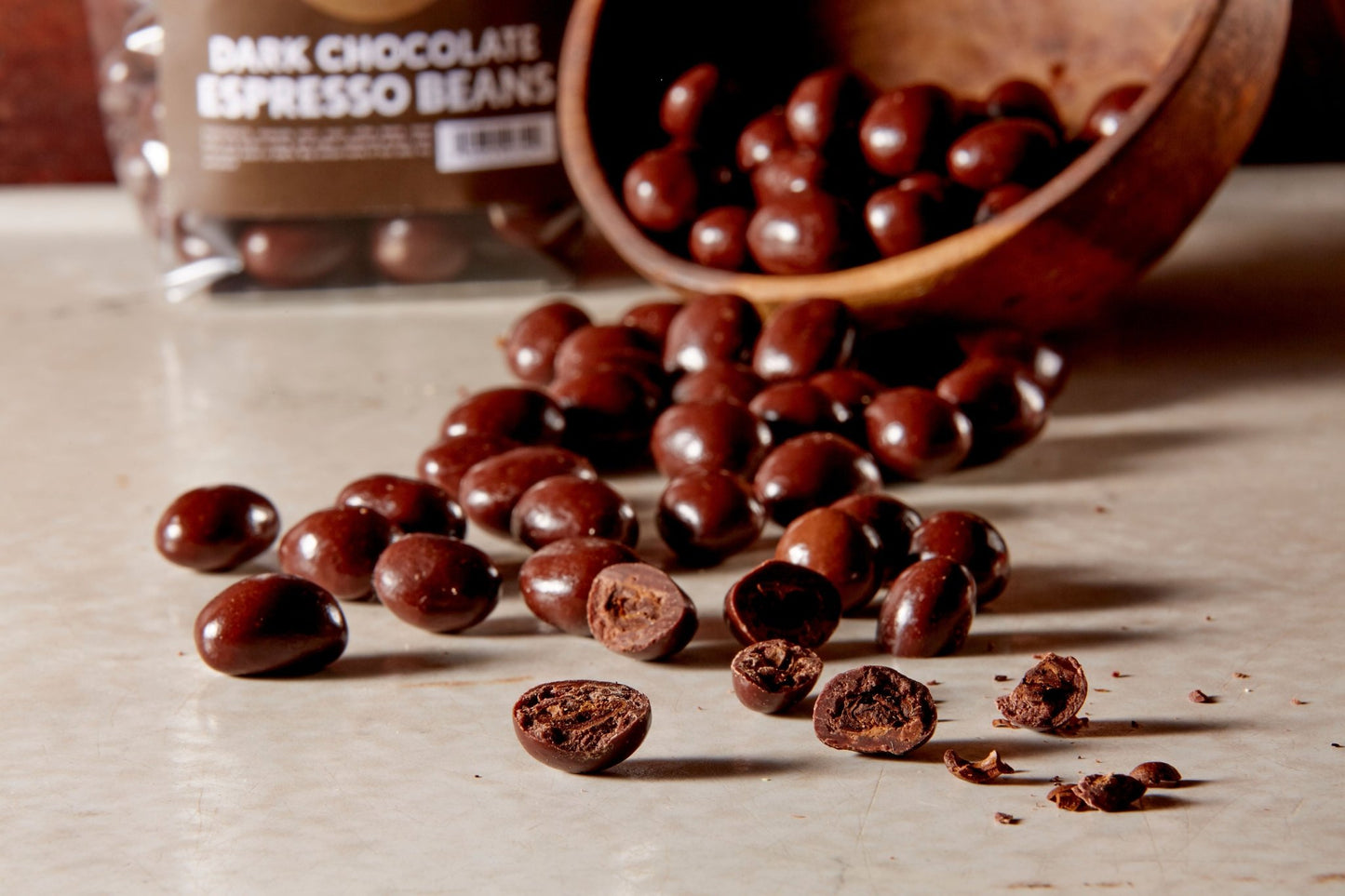 Dark chocolate espresso beans poured from a wooden bowl some broken open in the front, a branded bag full in the background