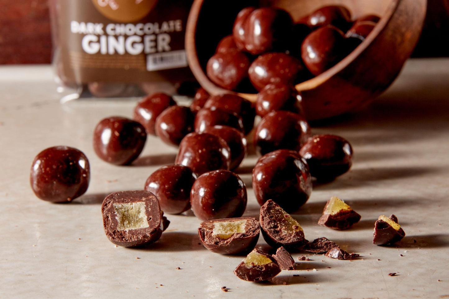Dark chocolate ginger poured from a wooden bowl some broken open in the front and a branded bag of them in the background